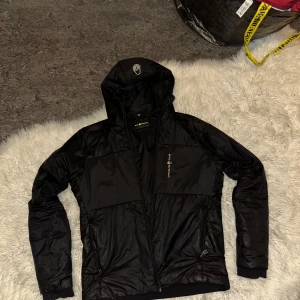 Svart pufferjacka från Sail Racing - Svart pufferjacka från Sail Racing med huva och dragkedja framtill. Jackan har två sidofickor med dragkedja och en bröstficka med Sail Racing-logga. Materialet är glansigt och syntetiskt. Snygg och sportig look med långärmad passform.