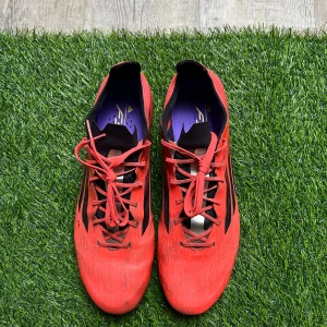 Röda Adidas F50 fotbollsskor - Säljer ett par röda Adidas F50 fotbollsskor med svarta detaljer och snörning. Skorna har rund tå och platt sula med dobbar för gräsplan. Ovandelen är tillverkad i syntetmaterial och har en sportig, smal siluett. Perfekt för dig som vill sticka ut på planen.