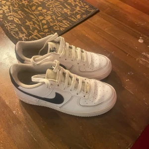 Nike Air Force 1 vita sneakers - Säljer ett par klassiska Nike Air Force 1 sneakers i vitt skinn med svart swoosh på sidan. Modellen har rund tå, snörning och platt sula med tydligt mönster undertill. Perfekt för dig som gillar stilrena och tidlösa sneakers med streetkänsla.