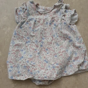 Babyklänning - Supersöt babyklänning från H&M i ekologisk bomull. Klänningen är vit med blommigt mönster i rosa, blått och gult, har volangärmar och praktiska knappar på axeln samt bodysuit med tryckknappar undertill.