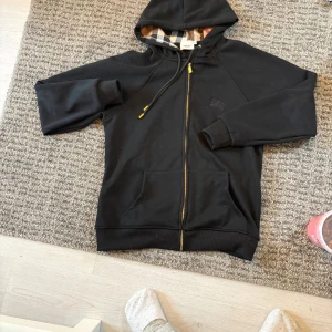  Burberry hoodie  - Svart hoodie från Burberry med klassiskt rutigt foder i huvan. Tröjan har guldfärgade detaljer på snörningen och dragkedja, samt två fickor framtill. Perfekt för dig som gillar stilrena och lyxiga streetwear-plagg. Jag har fått  in flera storlekar: S,M och L finns inne, frakt finns via Plick. Hör av er vid frågor.  