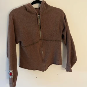 Brun hoodie med vingar från La Luna - Brun stickad hoodie med dragkedja och huva från La Luna. På ryggen finns ett stort broderat vingmotiv i beige och på ärmen sitter en patch med röd detalj. Tröjan har lång ärm och råa kanter för en chill vibe.