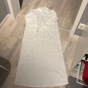 Vit långklänning från KappAhl - Enkel och stilren vit långklänning från KappAhl i storlek 158/164. Klänningen har lång ärm, hög hals och raka linjer. Perfekt för dig som gillar minimalistisk stil och vill ha något klassiskt och bekvämt.