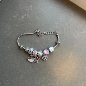 Silverfärgat armband med rosa berlocker - Snyggt armband i silverfärg med flera berlocker, bland annat ett glittrigt hjärta, en svan, en bokstav V med rosa stenar och en blomma. Armbandet har även pärlor i rosa och vita toner samt en justerbar kedja. Perfekt för dig som gillar personliga detaljer.