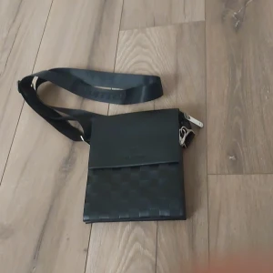 Svart axelväska med rutigt mönster - Snygg svart axelväska med rutigt mönster på framsidan och bred axelrem. Väskan har ett stilrent och modernt utseende, perfekt för att bära det viktigaste. Materialet ser ut att vara skinn eller syntet och väskan stängs med lock och dragkedja.