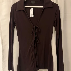 Brun långärmad topp från Gina Tricot - Säljer en brun långärmad topp från Gina Tricot i mjuk jersey. Toppen har en djup v-ringning, krage och snygga knytband framtill som ger en trendig look. Passformen är slim och materialet känns skönt mot huden. Perfekt att styla med jeans eller kjol.