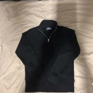 Svart stickad half zip från Jack & Jones - Svart stickad tröja från Jack & Jones med half zip-dragkedja och hög krage. Tröjan har långärmad design och är tillverkad i mjukt material som passar perfekt när det är lite kyligare. Enkel och stilren look som funkar till jeans eller chinos. Det finn defekt.