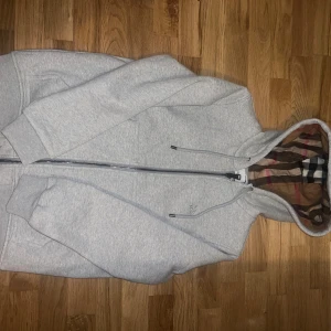 Grå Burberry hoodie - Snygg grå hoodie från Burberry med dragkedja och klassisk rutig insida i huvan. Tröjan har broderad Burberry-logga på bröstet, långa ärmar och ribbade muddar. Perfekt för dig som gillar stilrena detaljer och ikoniskt mönster. M men passar L