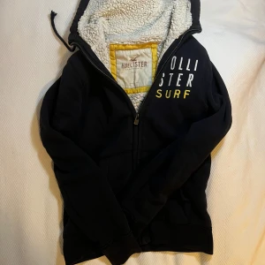 Svart zip hoodie från Hollister med foder - Svart hoodie från Hollister med broderad text i vitt och gult på bröstet. Insidan är fodrad med mjuk fuskpäls i vitt, vilket gör den extra mysig. Tröjan har huva med snörning och dragkedja framtill. Perfekt för kyliga dagar.