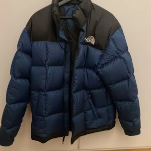 Blå och svart dunjacka The North Face - Säljer en blå och svart The North Face 1996 Lhotse dunjacka. Jackan är puffer-modell med stora quiltade sektioner, hög krage och dragkedja framtill. Ikonisk logga på bröstet och ryggen. Perfekt för kalla dagar och riktigt snygg streetstil.