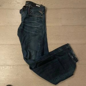 Replay Anbass mörkblå jeans - Snygga mörkblå Replay Anbass jeans med klassisk femficksdesign och diskreta slitningar. Jeansen har raka ben och normal passform, med detaljer som Replay-logga på bakfickan och läderpatch i midjan. Perfekta för dig som gillar stilrena och coola jeans. Längd 34 och midja 31