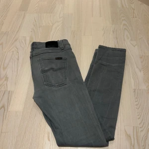 Grå jeans från Nudie Jeans, 31/34 - Snygga grå jeans i modellen LEAN DEAN från Nudie Jeans i klassisk femficksmodell. Byxorna har raka ben, svarta detaljer på bakfickan och ett svart läderpatch med logga i midjan. Materialet är mjukt jeans-tyg och passformen är normal. Perfekt för dig som gillar stilrena och enkla jeans.