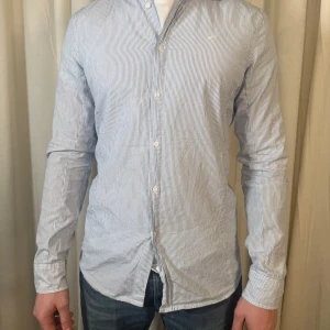 Randig skjorta från Diesel, storlek L - Snygg skjorta från Diesel i storlek L med tunna blå och vita ränder. Skjortan har klassisk krage, långa ärmar och knappar framtill. Tillverkad i 100% bomull för en skön och lätt känsla. Perfekt för dig som gillar stilrena och fräscha plagg.