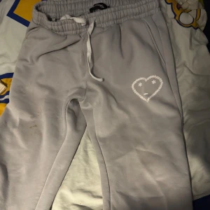 Grå mjukisbyxor med hjärtdetalj - Grå mjukisbyxor med resår och snörning i midjan. På vänster ben finns ett vitt hjärta med ansikte som tryck. Byxorna har avslappnad passform och är perfekta för chill dagar. Mjukt material som känns skönt mot huden.