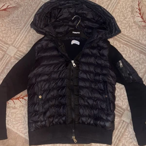 Svart Moncler cardigan med huva - Svart cardigan från Moncler med stickade ärmar och detaljerade fickor med dragkedja. Jackan har en stor huva, ribbade muddar och quiltad front i syntetmaterial. Moncler-logga på ärmen och coolt serietryck på insidan. Perfekt för dig som gillar stil och premium. NFC taggen funkar som man kan se på sista bilden. Jackan är i storlek S. Jackan sitter relativt slim vilket gör att jackan inte ser hängig ut när man har på sig den. Priset är ej hugget i sten så tveka inte att fråga!