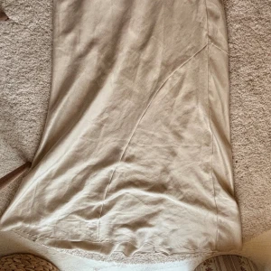 Beige långkjol i satin med dragkedja - Säljer en beige långkjol i mjukt satintyg med snyggt snitt och diskret dragkedja i sidan. Kjolen har en lätt glansig finish och faller snyggt längs benen. Perfekt för dig som gillar minimalistisk och clean stil.