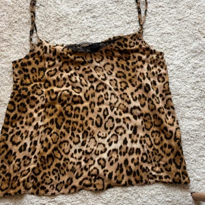 Leopardmönstrad linnetopp med spets - Säljer en leopardmönstrad linnetopp med tunna axelband och svart spetsdetalj vid urringningen. Toppen har en glansig finish och är i ett mjukt, följsamt material som känns lyxigt. Perfekt för dig som vill sticka ut med ett djurmönster och snygga detaljer.
