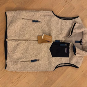 Vit fleeceväst från Patagonia - Vit fleeceväst från Patagonia med marinblå detaljer och bröstficka. Västen har hel dragkedja, två sidofickor med dragkedja och meshfoder på insidan. Perfekt för lager-på-lager och outdoor vibes. Storlek L sitter som en M slim fit 