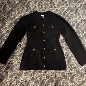 Svart kofta med guldknappar från H&M - Snygg svart kofta från H&M med guldiga knappar framtill och på fickorna. Modellen är långärmad och har två stora fickor framtill. Koftan är stickad och har en klassisk, lite längre passform som ger en stilren look. Koftan är i storlek XS men passar även de som bär S om de vill ha en figurnära look. (Jag på bilden bär vanligtvis S). Använd vid ett tillfälle. 