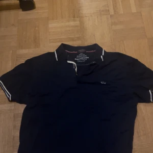 Marinblå pikétröja från Paul & Shark - Snygg marinblå pikétröja från Paul & Shark med klassisk krage och vita detaljer på krage och ärmslut. Tröjan har korta ärmar och liten logga på bröstet. Perfekt för en stilren och sportig look.