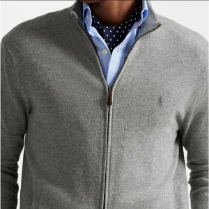 Ullkofta Polo Ralph Lauren - Snygg gråmelerad kofta från Polo Ralph Lauren i mjuk ull. Full zip framtill, ribbade muddar och diskret broderad logga på bröstet. Perfekt att slänga på över en skjorta för en clean och stilren look. Ny pris är 2 999kr