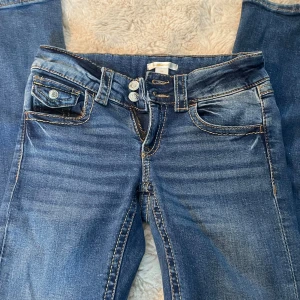 Blå jeans från Gina Tricot - Snygga blå jeans från Gina Tricot med klassisk femficksdesign och kontrastsömmar. Modellen har dubbla knappar i midjan och en liten ficka med lock framtill. Jeansen är bootcut och lågmidjade.
