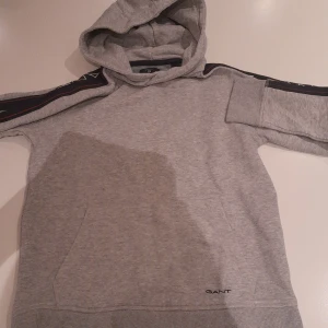 Grå hoodie från Gant med huva - Säljer en grå hoodie från Gant med huva och svarta ränder med logga längs ärmarna. Tröjan har känguruficka framtill och ribbade muddar. Perfekt för chill dagar och snygg till jeans eller mjukisbyxor.