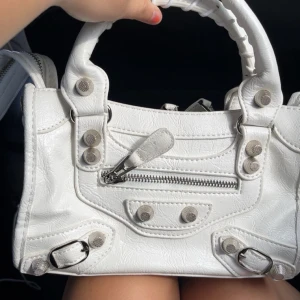 Väska med nitar - Snygg vit handväska i skinnimitation med coola silverfärgade detaljer och dragkedja framtill. Väskan har ett unikt designat handtag och flera dekorativa nitar. Perfekt storlek för att få plats med det viktigaste och passar till många olika stilar.