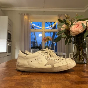 Golden Goose Superstar vita sneakers - Golden Goose Superstar sneakers i vitt skinn med grå stjärndetalj på sidan och klassisk snörning. Platt sula och rund tå. Snyggt slitna detaljer för en cool och avslappnad look. Perfekta för dig som gillar streetstyle och vill ha något unikt. Tror de är storlek 41 men de passar folk som bär 43