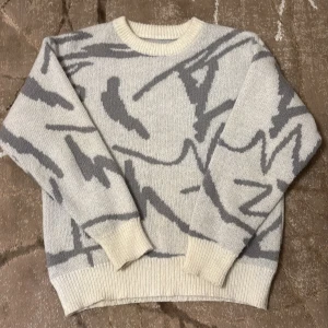 Gråvit stickad tröja med mönster - Säljer en gråvit stickad tröja med abstrakt mönster i grått. Tröjan har rund hals, ribbade muddar och långärmad passform. Perfekt för dig som gillar en chill och stilren look med lite edge.