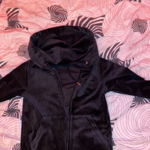 Svart velourjacka Juicy Couture S - Svart velourjacka från Juicy Couture i storlek S. Jackan har huva med snörning, dragkedja framtill och två fickor. Mjuk och glansig yta med diskret logga på bröstet. Perfekt för en chill nästan oanvänd 
