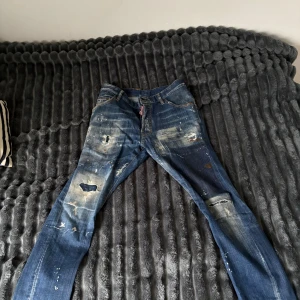 Blå distressed jeans från Dsquared2 - Säljer ett par blå jeans från Dsquared2 med coolt distressed och slitna detaljer, färgstänk och patchade partier. Klassisk femficksmodell med DSQ2-logga på bakfickan och röd DSQUARED2-tag vid gylfen. Snygga kontrastsömmar och rak passform.