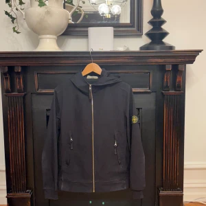 Svart vindjacka från Stone Island - Svart vindjacka från Stone Island med huva och klassisk logotyp-patch på vänster ärm. Jackan har två fickor med dragkedja framtill och hel dragkedja. Materialet är mjukt och lätt, perfekt för dig som gillar streetwear och snygga detaljer. Lite slitningar längst sömmarna och även vid armbågarna annars är den i ok skick.