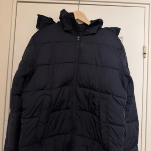 Svart pufferjacka med huva från dressman - Säljer en svart pufferjacka med huva och dragkedja framtill. Jackan har två sidofickor med dragkedja och är quiltad för extra värme. Perfekt för kalla dagar och har en stilren look som funkar till det mesta. Nypris 800kr, använd typ 2 vintrar