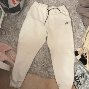 Beiga mjukisbyxor från Nike XL - Säljer ett par beiga mjukisbyxor från Nike i storlek XL. Byxorna har svart snörning i midjan, elastisk midja och ribbade muddar vid bensluten. Nike-logga i svart på vänster lår. Sköna och stilrena, perfekta för chill eller träning.