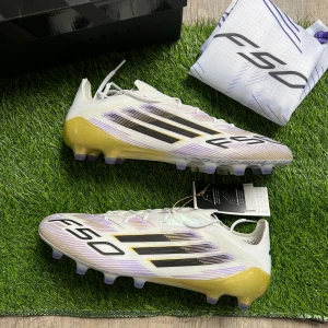 Adidas F50 fotbollsskor vit/guld - NYA - Säljer ett par Adidas F50 fotbollsskor i vitt med guldiga och svarta detaljer samt lila inslag. Skorna har snörning, platt sula med dobbar och är tillverkade i syntetmaterial för lätthet och snabbhet på planen. Snyggt randigt mönster och medföljer F50-påse.