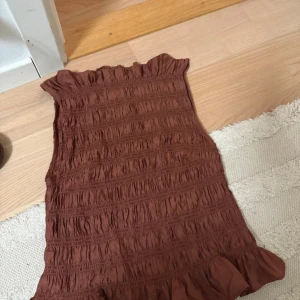 Brun kort volangklänning i bomull - Supersöt brun kort klänning med volangkant nertill och smockad struktur över hela plagget. Ärmlös modell med lätt rynk vid halsringningen. Perfekt för varma dagar och enkel att styla med sneakers eller sandaler.