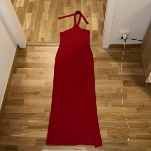 Röd långklänning med slits - Är 1,69 lång och den är pitelite kort Snygg röd långklänning med halterneck och slits framtill. Klänningen har en tight passform och är ärmlös med knytband runt nacken. Perfekt för dig som vill sticka ut med en enkel men elegant look.