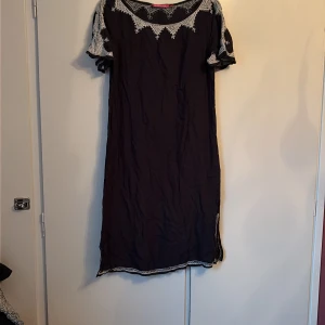 Svart lklänning med spets från Indiska - Svart klänning från Indiska i storlek M med vita spetsdetaljer vid halsringning, ärmar och nederkant. Klänningen har korta ärmar och en slits på sidan. Materialet känns lätt och luftigt, perfekt för dig som gillar boho-vibe och snygga kontraster.