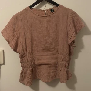 Beige blus med volang från SHEIN - Säljer en beige blus från SHEIN i storlek M. Blusen har korta ärmar, rund hals och volangdetaljer vid midjan och nederkant. Materialet är mjukt och strukturerat, vilket ger en chill och luftig känsla. Perfekt för dig som gillar en enkel men söt look.