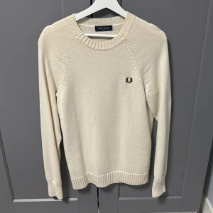 Beige stickad tröja från Fred Perry - Snygg beige stickad tröja från Fred Perry med rund halsringning och diskret broderad logga på bröstet. Tröjan har ribbade muddar och är långärmad, perfekt för lager-på-lager under hösten och vintern. Ett av dem skönaste och varma tröjorna jag haft på mig synd att den inte längre passar mig.