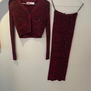 Vinröd ribbad långkjol med långärmad axel vadderad toppfrån Portrait - Säljer en vinröd långkjol från Portrait med svart ribbat mönster och knappar längs hela framsidan. Kjolen har en figurnära passform och är tillverkad i ett stretchigt, stickat material som känns mjukt mot huden. Perfekt för dig som gillar en stilren men edgy look.