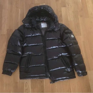 Svart Moncler pufferjacka med huva - Svart pufferjacka från Moncler med glansig finish och huva. Jackan har Moncler-logga på ärmen, två sidofickor med dragkedja och quiltad design. Insidan har ett serietidningsmärke och etikett. Perfekt för kalla dagar och riktigt snygg streetstil.