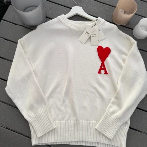 Vit stickad tröja från Ami Paris - Vit stickad tröja från Ami Paris med röd hjärtlogga och bokstaven A på bröstet. Tröjan har ribbade muddar vid ärmslut och nederkant samt rund hals. Materialet är mjuk ull och passformen är normal. Perfekt för dig som gillar stilrena detaljer och snygga loggor.