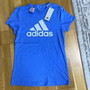 Blå Adidas t-shirt med logga - Blå t-shirt från Adidas med stor vit logga framtill. Klassisk rund hals och korta ärmar. Tillverkad i mjuk bomull för skön känsla. Perfekt för dig som gillar sportig och enkel stil.