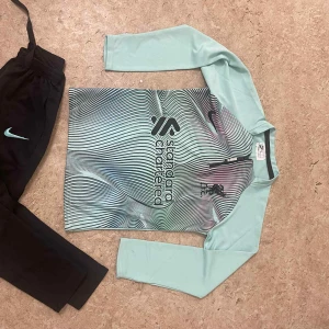 Liverpool långärmad fotbollströja Nike - Snygg långärmad Liverpool fotbollströja från Nike i ljusblått med vågigt randigt mönster på framsidan. Tröjan har tryckt Liverpool FC-logga och sponsortryck, samt rund hals. Materialet är lätt och andas, perfekt för träning eller match.