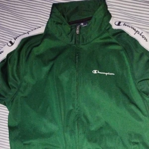 Grön vindjacka från Champion S - Grön vindjacka från Champion i storlek small. Jackan har dragkedja framtill, långärmade ärmar med vit logoband längs axlarna och Champion-tryck på bröstet. Materialet är polyester och jackan har en sportig, normal passform.
