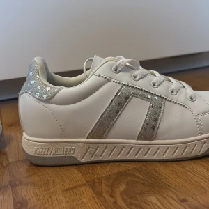 Vita Breezy Rollers sneakers med glitter - Snygga vita sneakers från Breezy Rollers med silvriga glitterdetaljer och stjärnor på hälen. Skorna är som rullator och har hjulen med sig men man kan använda dem utan hjulen också. Skorna har snörning och en klassisk rund tå. Perfekta för dig som vill sticka ut lite extra med glittriga detaljer. Tillverkade i syntetmaterial.