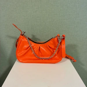 Orange axelväska från Mango med kedja - Snygg orange axelväska från Mango med en silverfärgad kedja som detalj. Väskan har en mjuk, rundad form och är gjord i glansigt syntetmaterial. Dragkedja upptill och justerbar axelrem. Perfekt för att lyfta din outfit med färg.