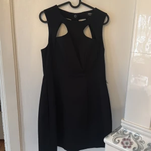 Svart cut-out klänning från Oasis - Svart kortklänning från Oasis med coola cut-out detaljer fram och bak. Klänningen är ärmlös och har en stilren, figurnära passform. Materialet är strukturerat och känns lite tjockare, perfekt för dig som gillar minimalistisk och edgy stil.